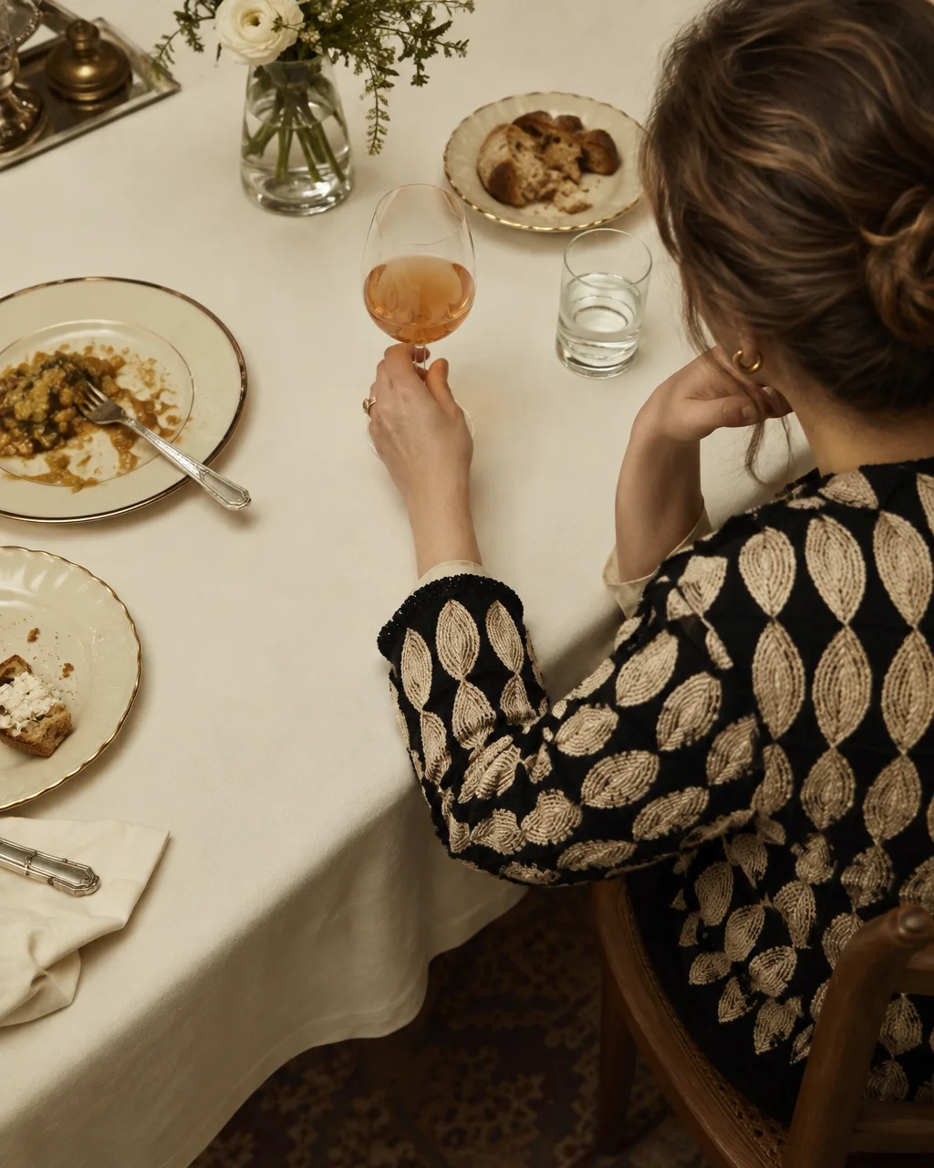 A woman in an embroidered jacket sits at an elegant dinner table holding a glass of rosé wine beside finished plates.