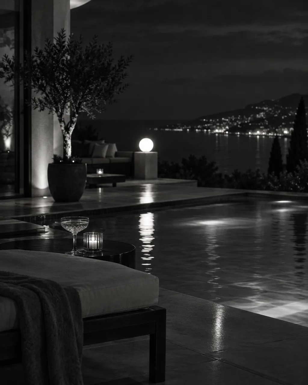 A monochrome night view across a pool terrace, with a cocktail glass and candle on a side table overlooking distant coastal lights.