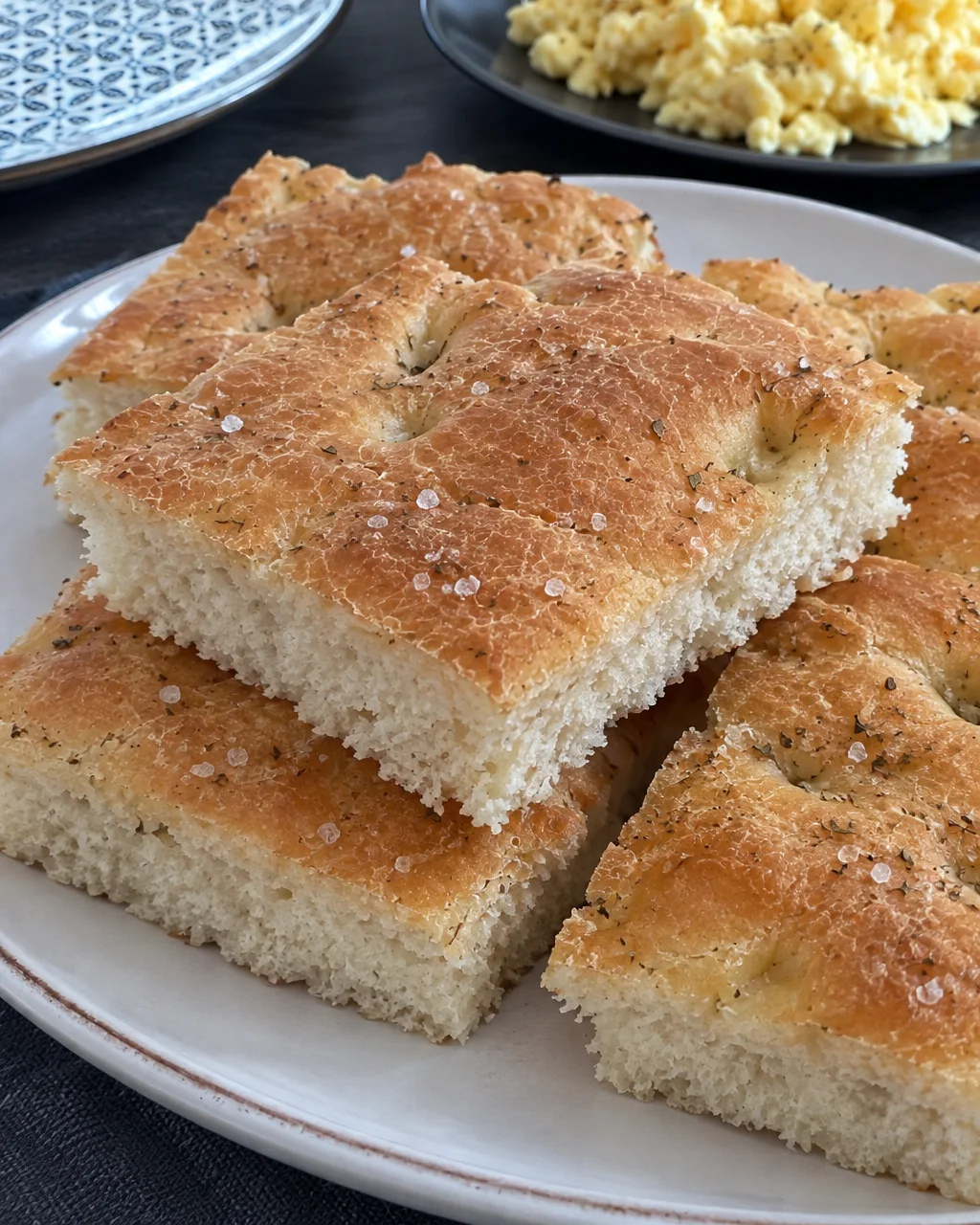 Golden squares of freshly baked focaccia topped with coarse salt and dried herbs, stacked on a white ceramic serving plate.