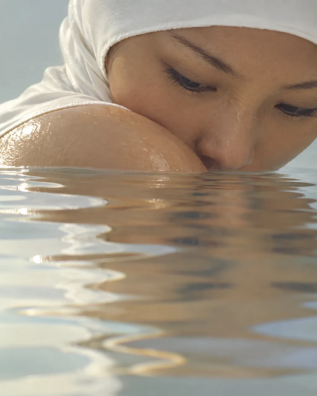 A close-up of a woman wearing a white headscarf resting near the surface of calm water, mirrored in gentle ripples.