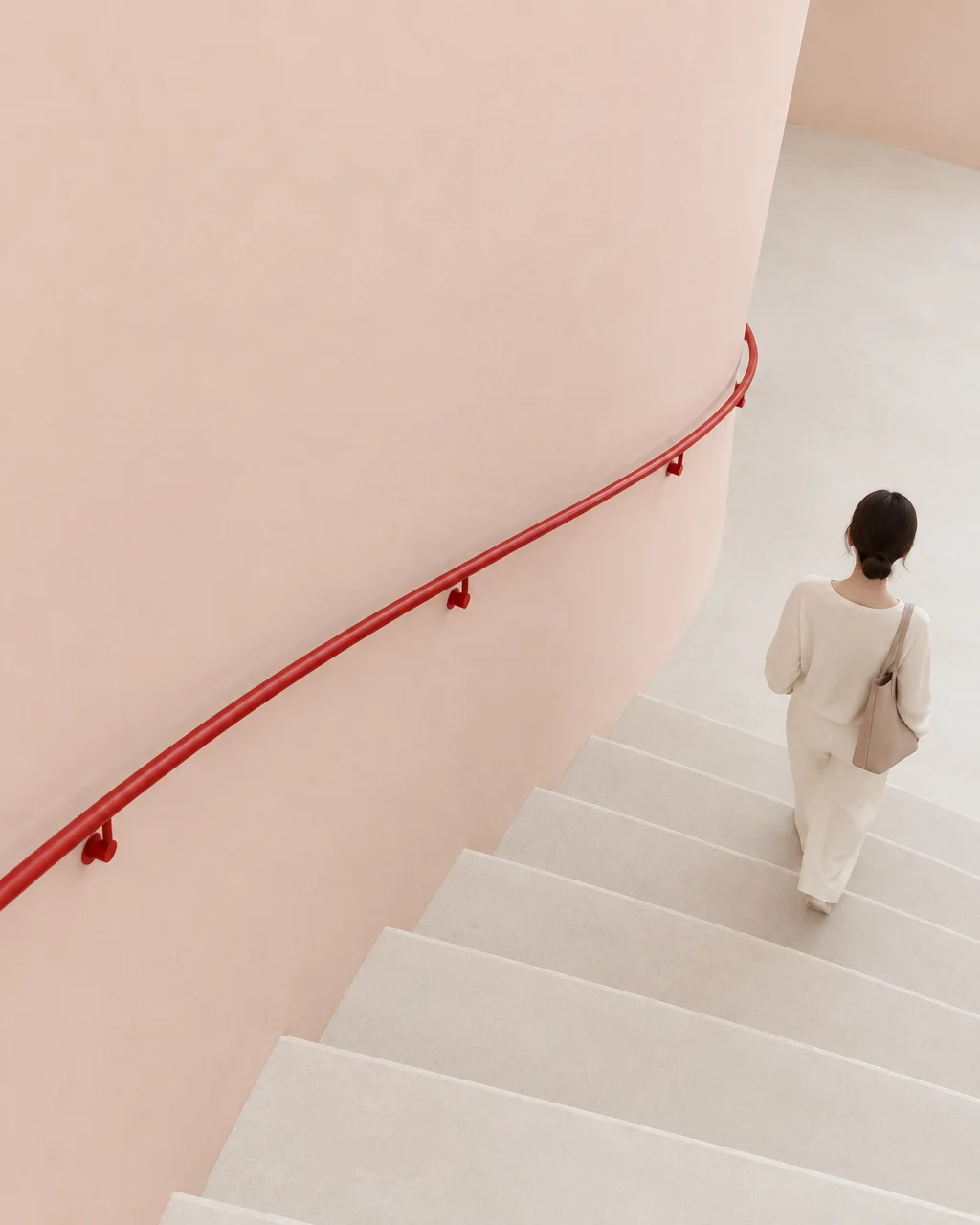A woman dressed in cream descends a minimalist concrete staircase alongside a curved blush wall with a striking red handrail.