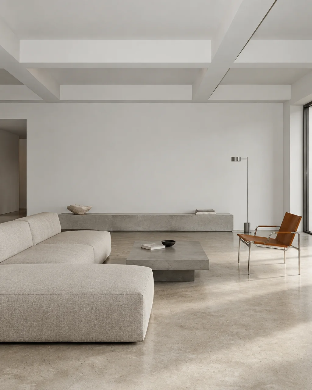 A minimalist living room featuring a low linen sectional, concrete coffee table, and cognac leather chair on polished concrete floors.