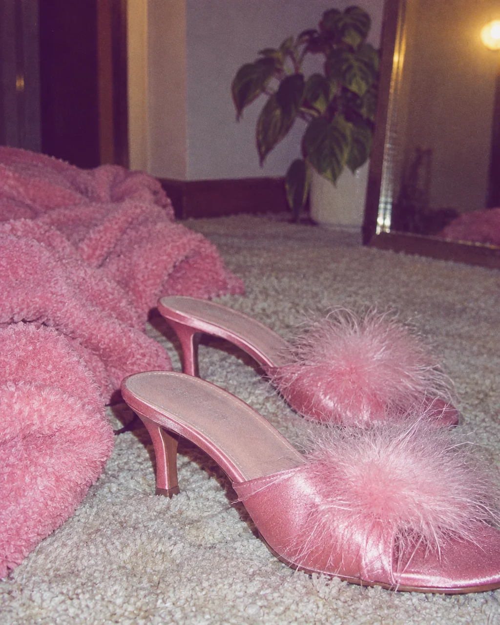 A pair of pink satin kitten heels with fluffy feather accents rests on carpet alongside a plush pink blanket.