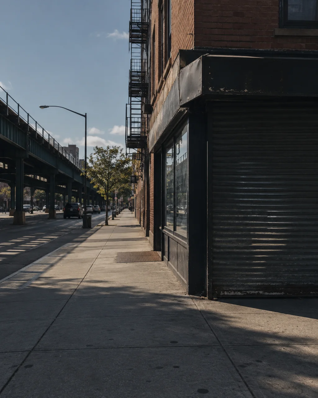 A quiet city sidewalk beside a brick storefront with a lowered metal shutter, running parallel to an elevated train track in angled sunlight.