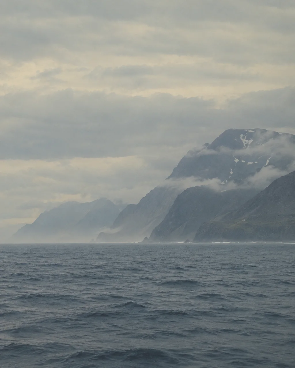 A dark, rippling ocean stretches toward rugged coastal mountains veiled in low clouds and sea mist under an overcast sky.