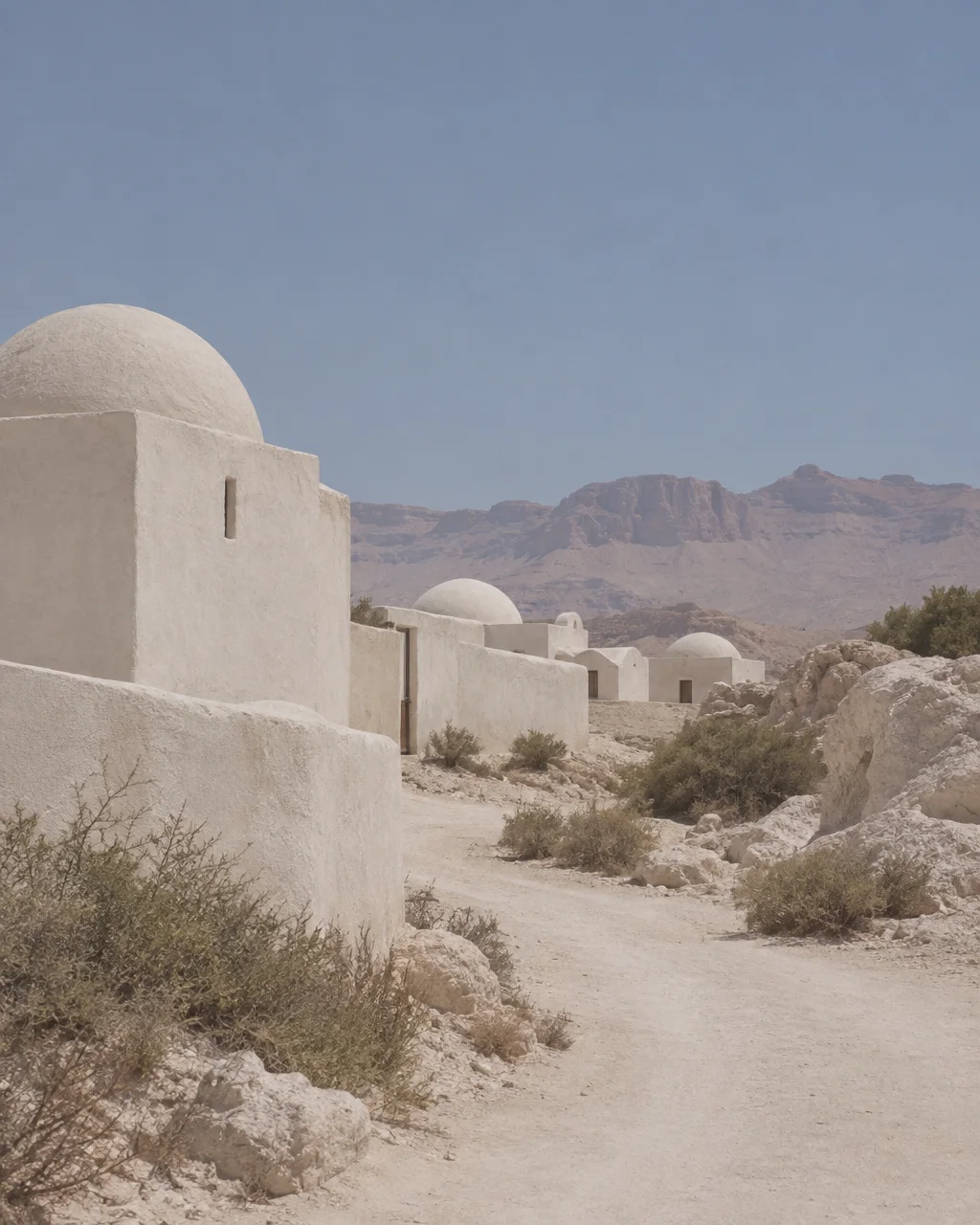 Whitewashed domed structures line a dusty track against rugged arid mountains under a pale sky.