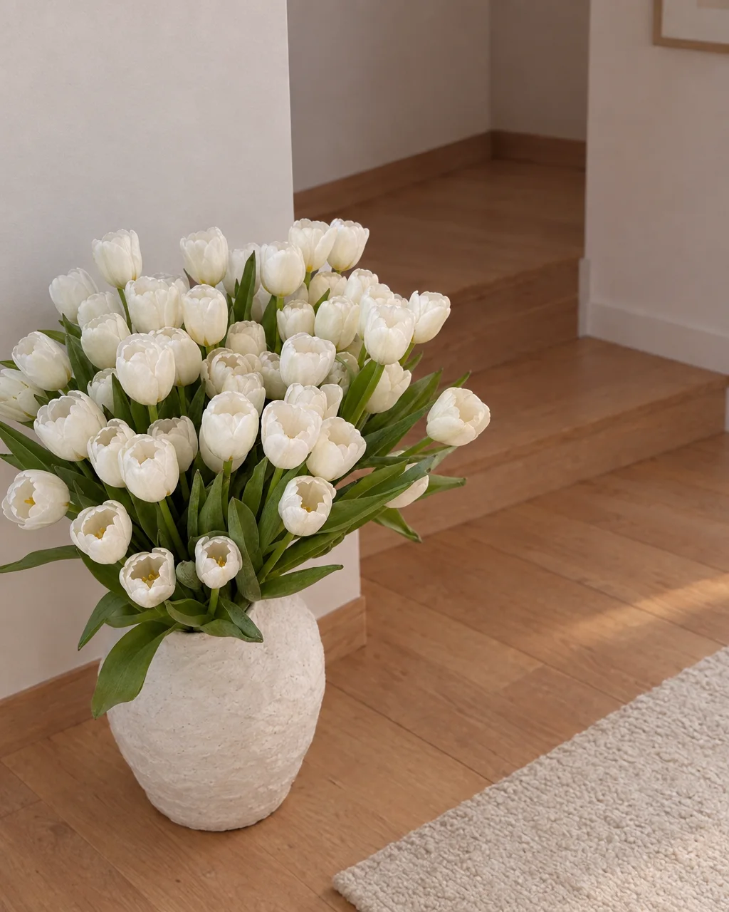 A large bouquet of fresh white tulips in a textured ceramic vase sitting on a light wooden floor in a serene interior.