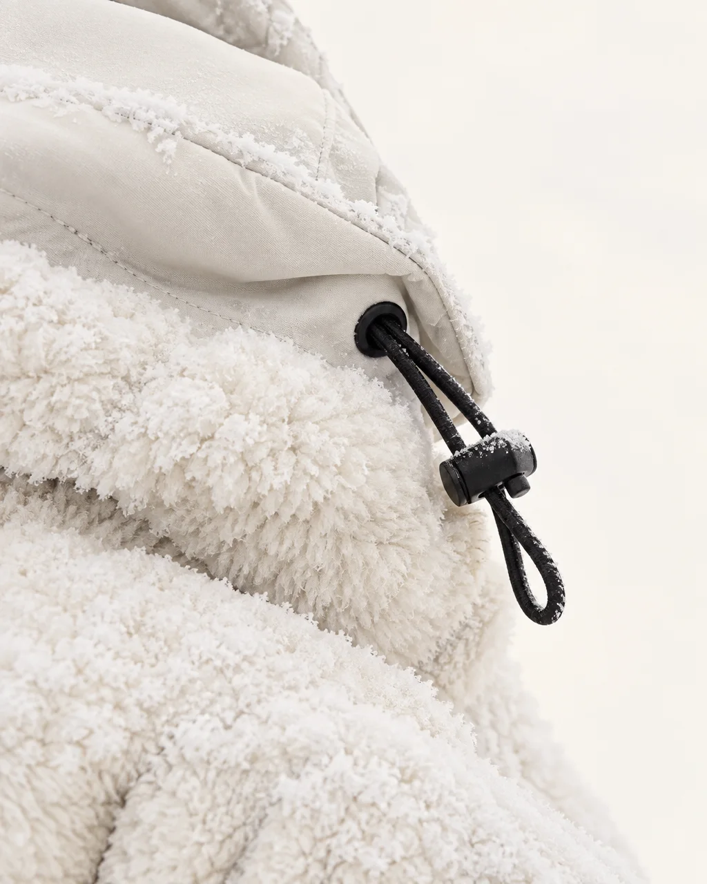 A macro detail of a white textured fleece jacket with a drawstring toggle lightly dusted in frost against a white backdrop.