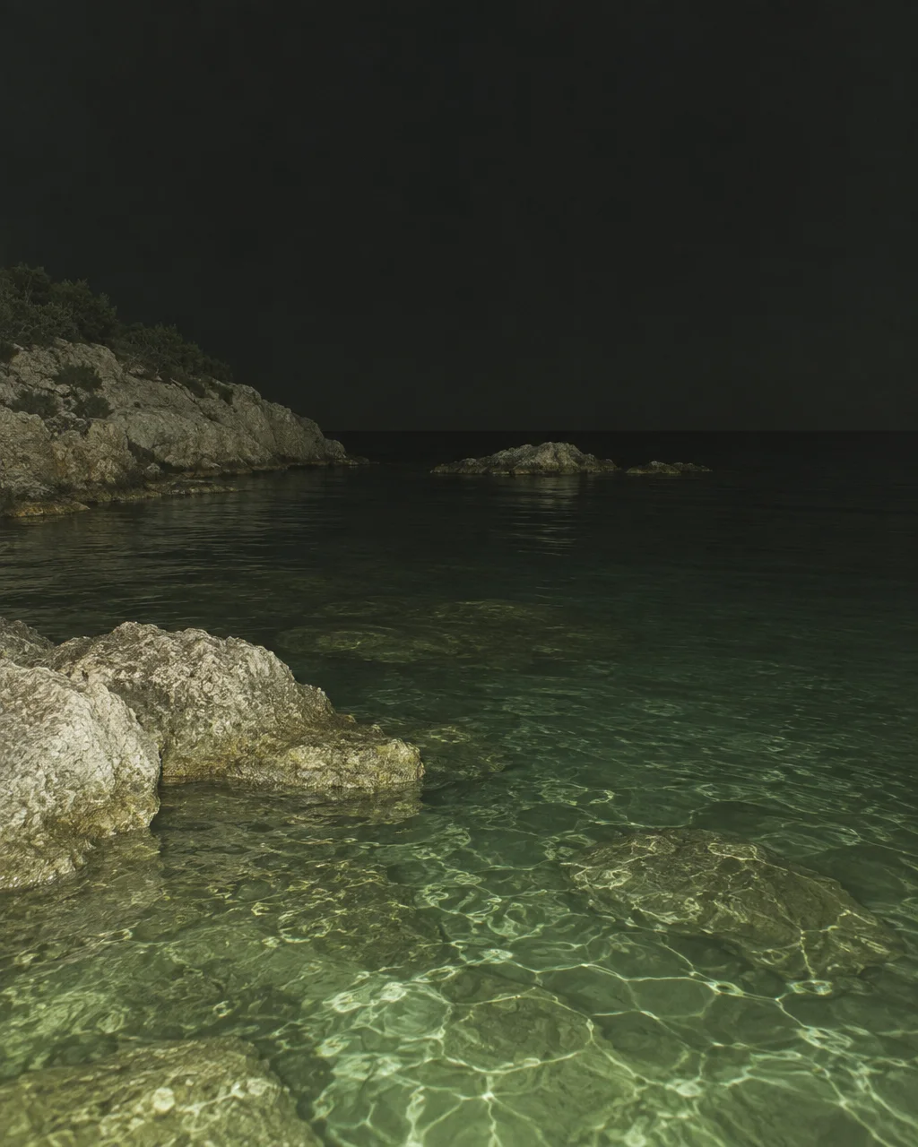 Clear turquoise water laps against rugged rocks along a quiet coastline illuminated in the darkness of night.