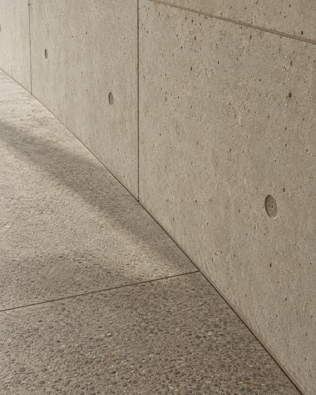 A smooth cast concrete wall with tie-rod marks meets a textured aggregate floor along a sharp diagonal seam.