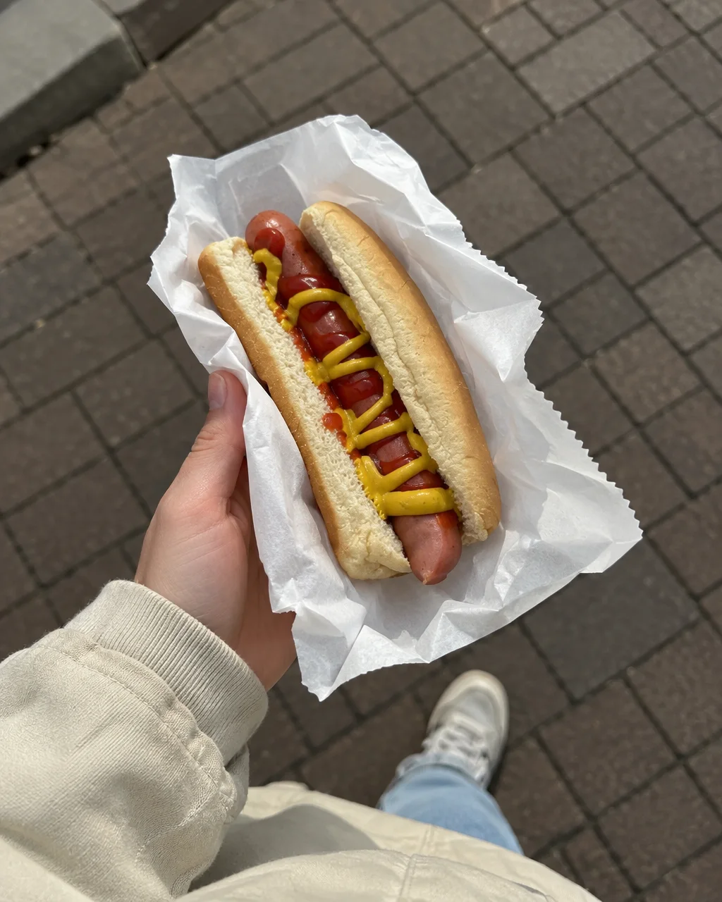 A hot dog topped with ketchup and mustard is held in a white paper wrapper over a paved sidewalk.