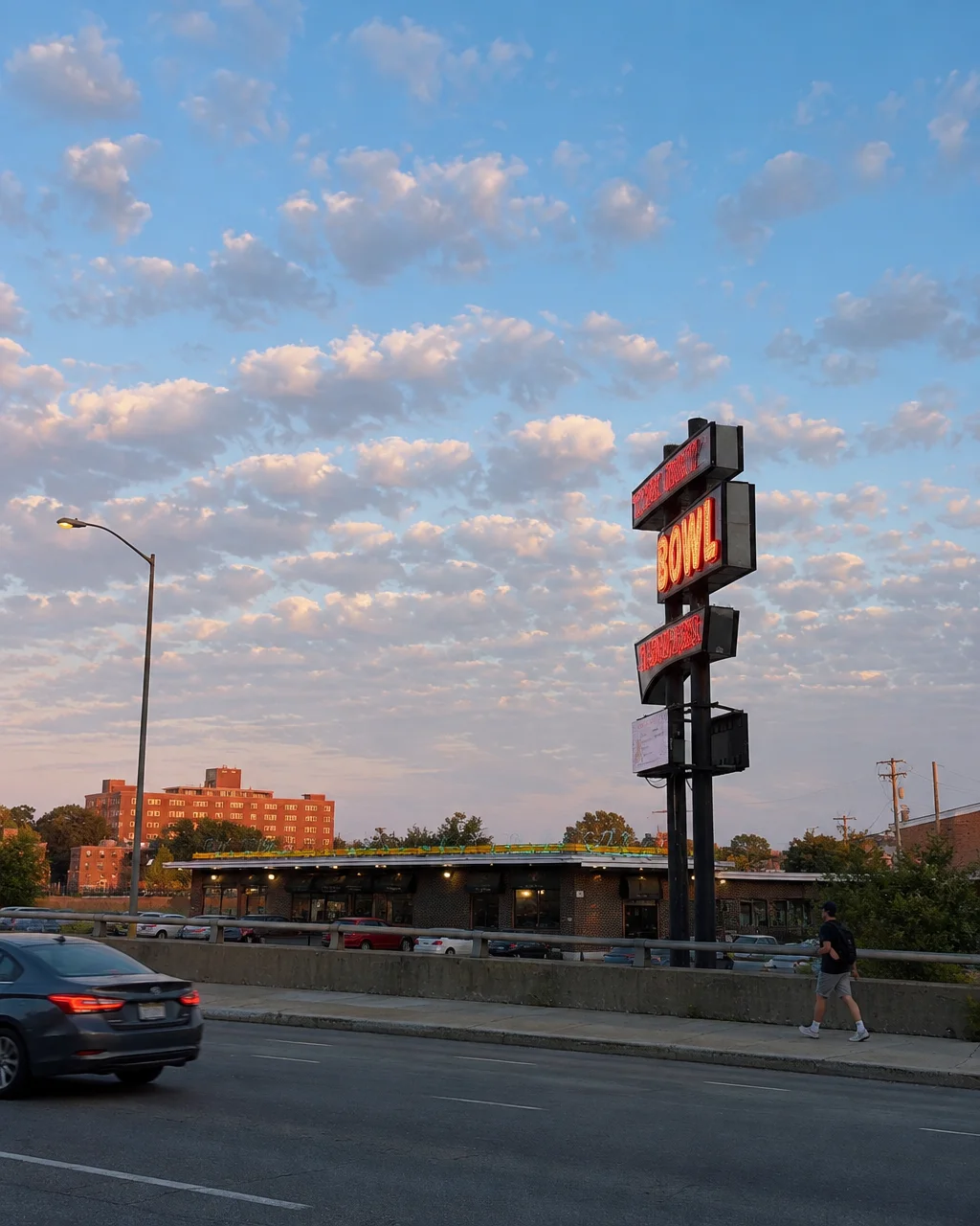 A tall vintage neon sign stands by a roadside under a soft cloudy evening sky with passing traffic and a pedestrian.