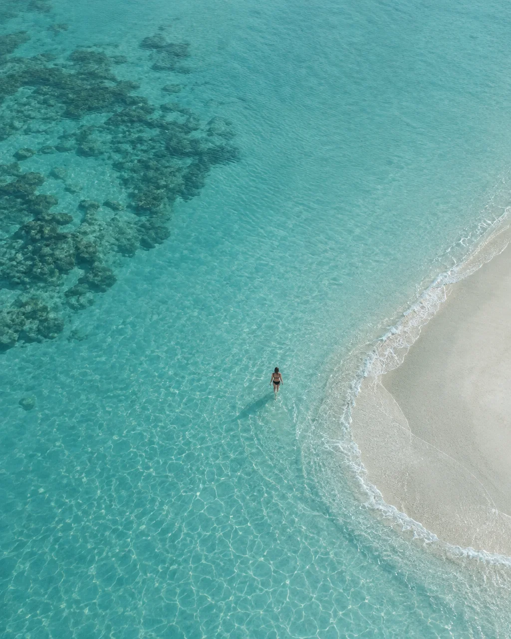 A lone person wades through transparent turquoise ocean water along the curve of a white sandbank and coral reef.
