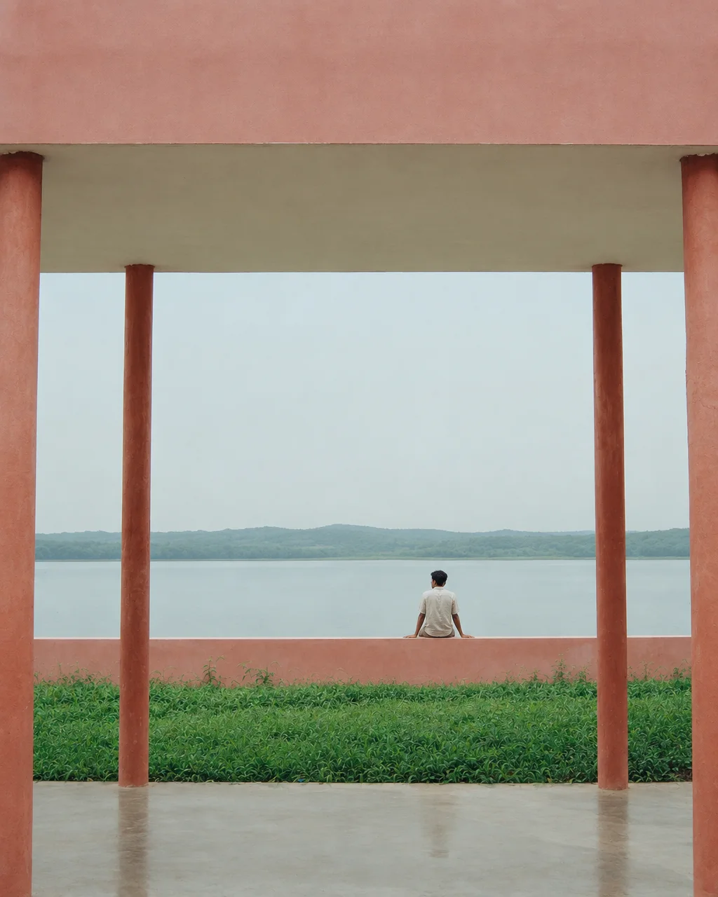 A person sits on a low terracotta wall looking out across a calm lake, symmetrically framed by structural pavilion pillars.