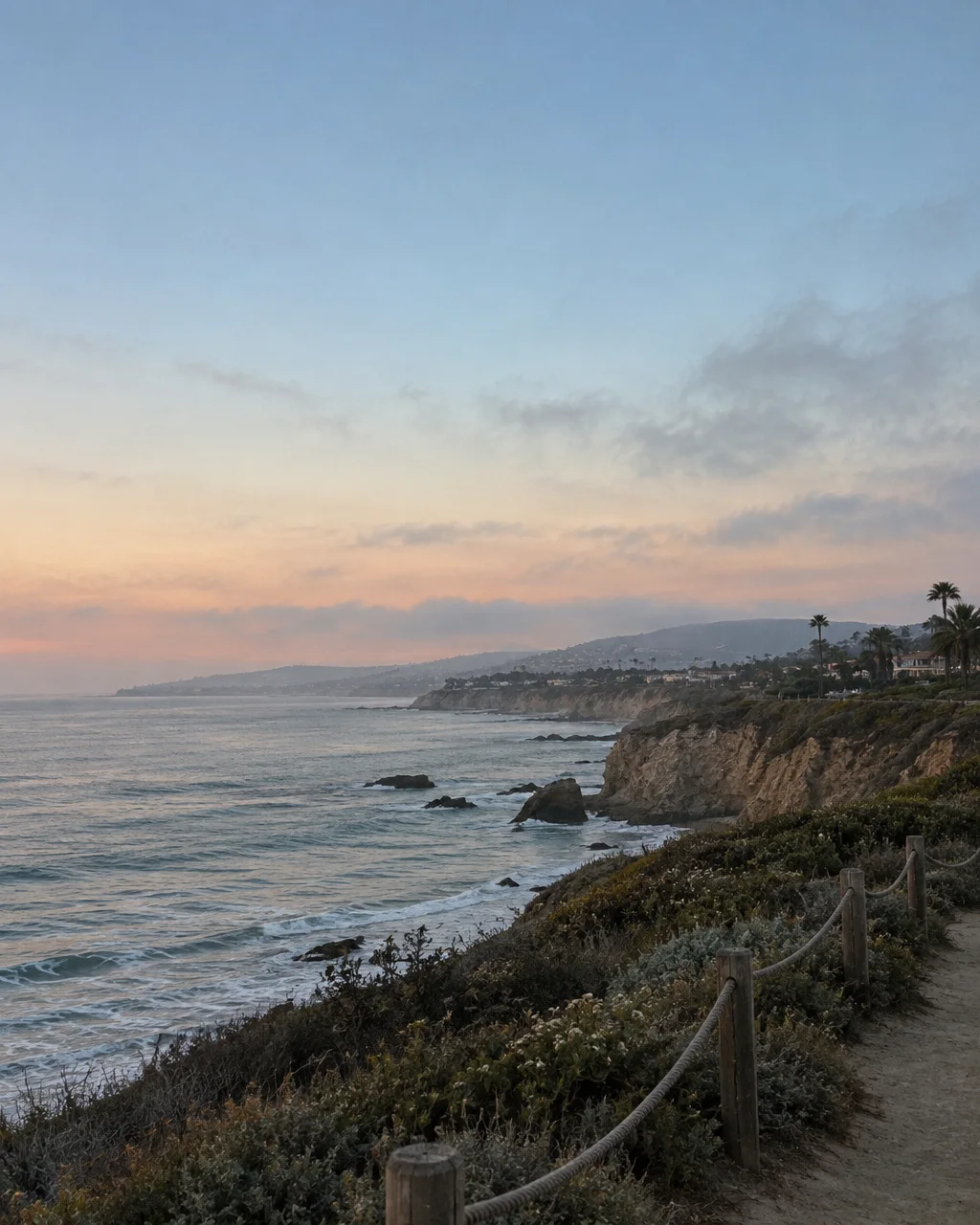 A coastal footpath with a rope fence overlooking rugged ocean cliffs under a soft pastel sunset sky.