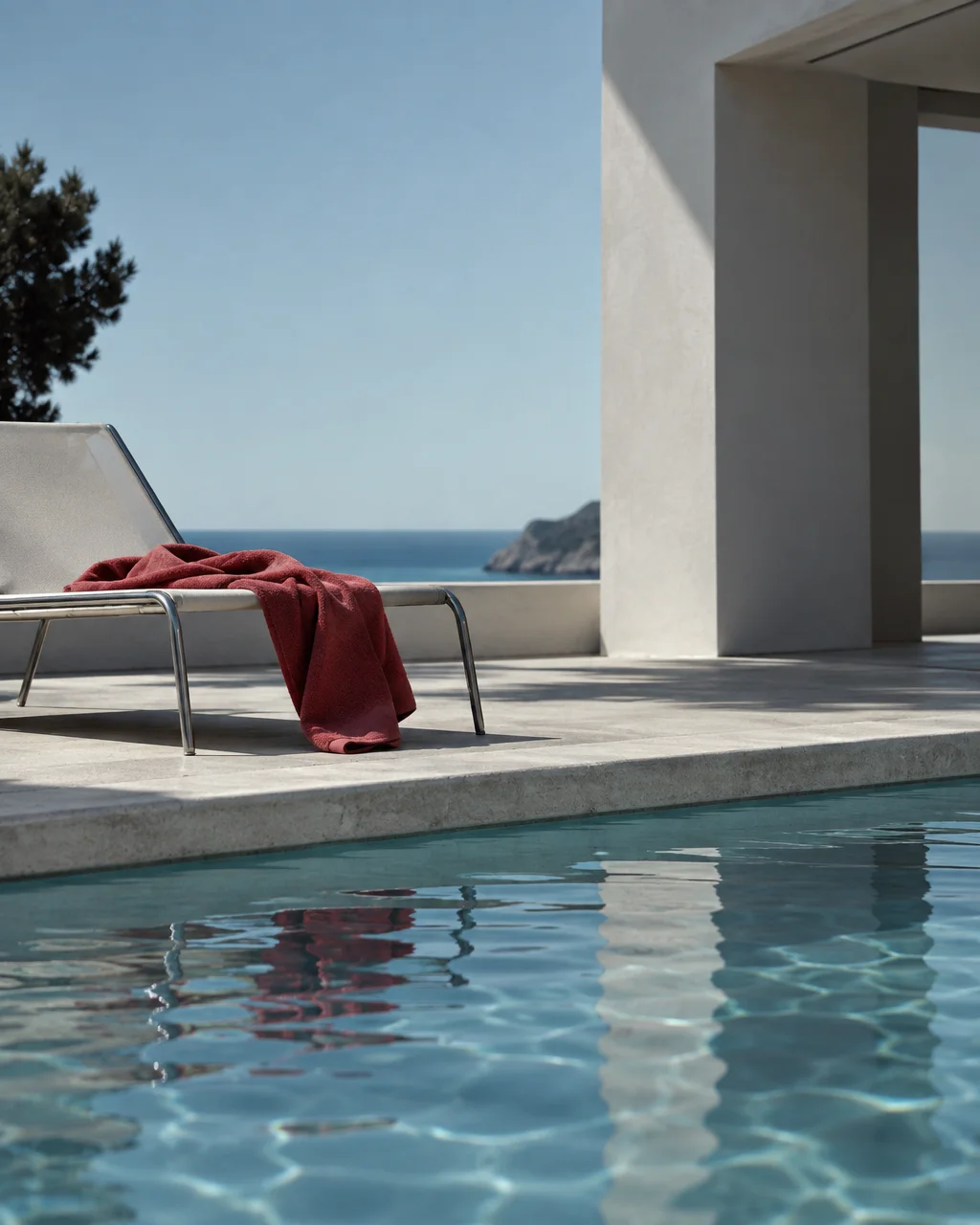 A red towel draped over a modern sun lounger beside a swimming pool, overlooking a calm coastal horizon.