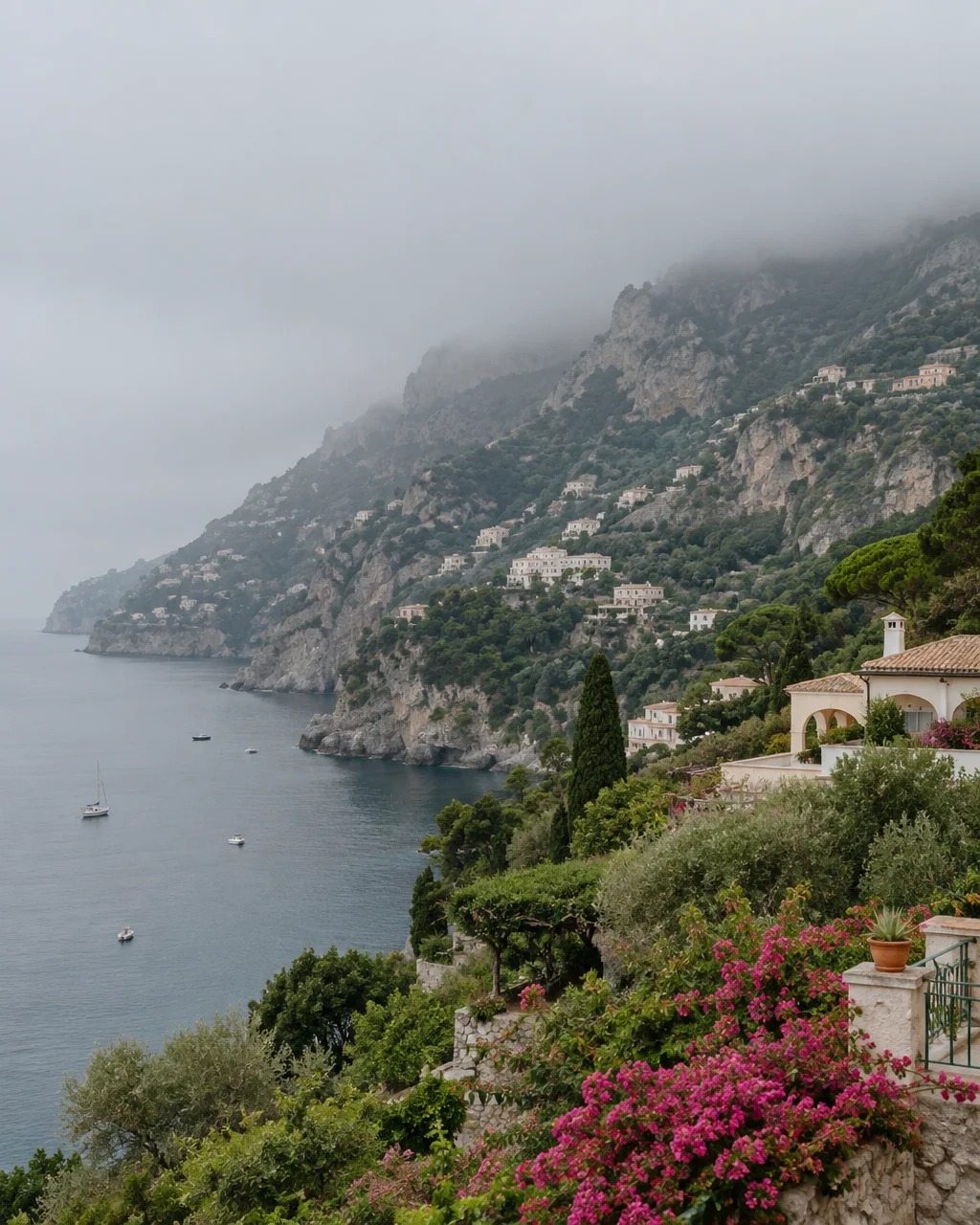 Villas and lush gardens cascade down steep rocky cliffs into a calm sea under a low, misty sky.