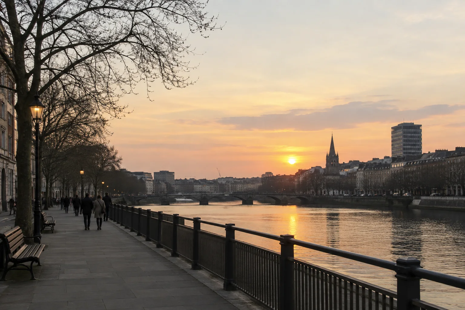 Pedestrians stroll along a wide paved riverside promenade lined with trees and lampposts under a warm sunset sky.