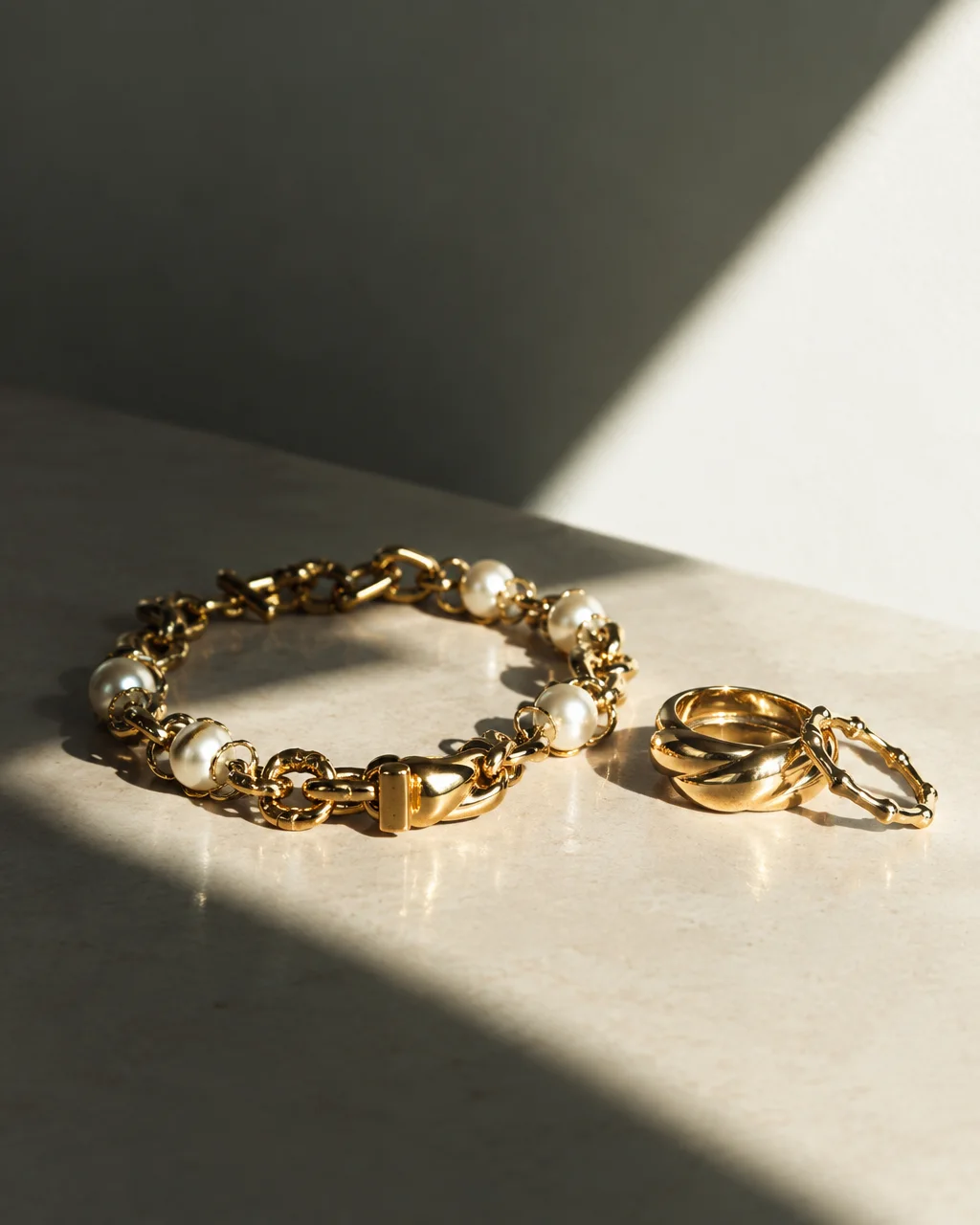 A heavy gold chain bracelet set with pearls resting beside gold rings on a stone surface under sharp diagonal sunlight.