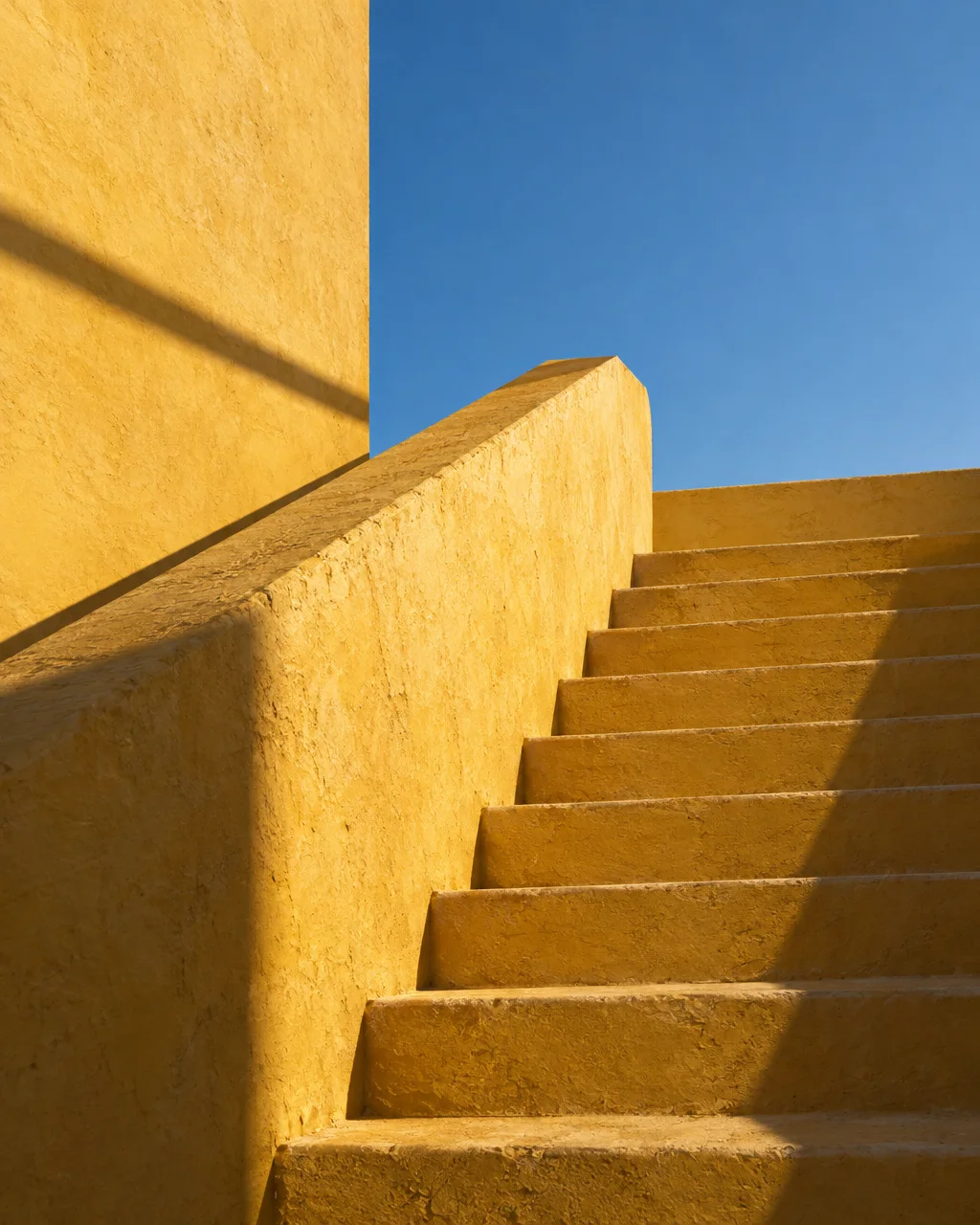 A textured yellow plaster staircase climbs toward a clear blue sky, casting crisp geometric shadows in bright sunlight.