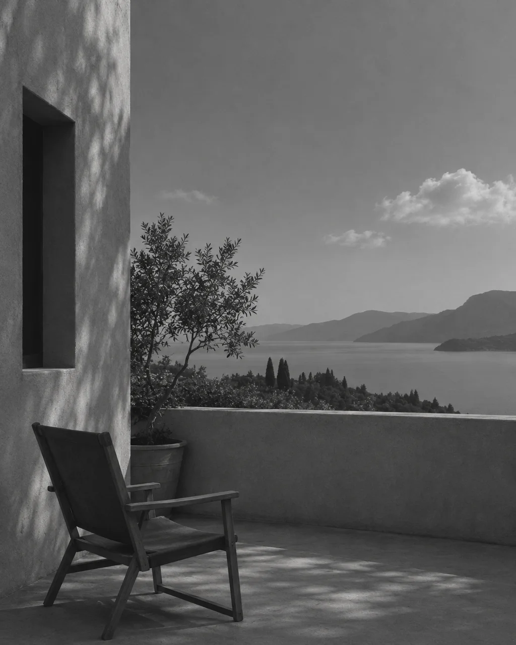 A wooden armchair rests in dappled shade on a quiet stone terrace overlooking a calm coastal bay and distant mountains in black and white.