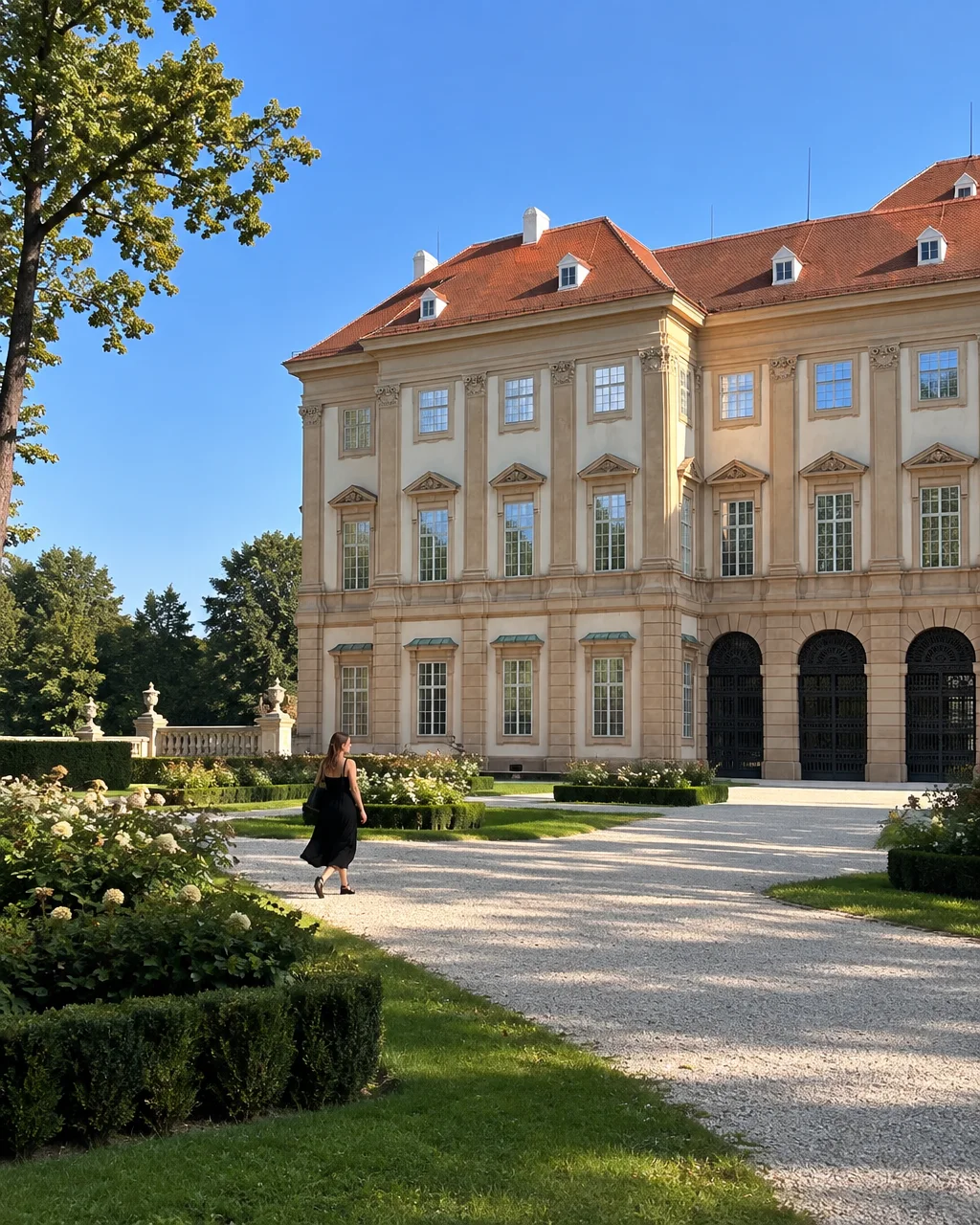 A woman in a black dress walks along a gravel path through manicured formal gardens beside a grand classical estate in bright daylight.