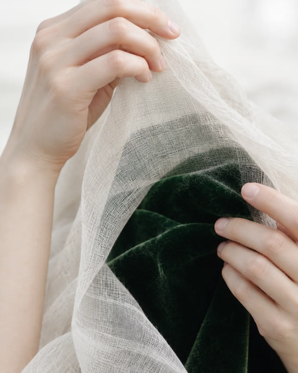 Hands lifting a sheer off-white open-weave fabric to reveal a deep emerald green velvet layer beneath.