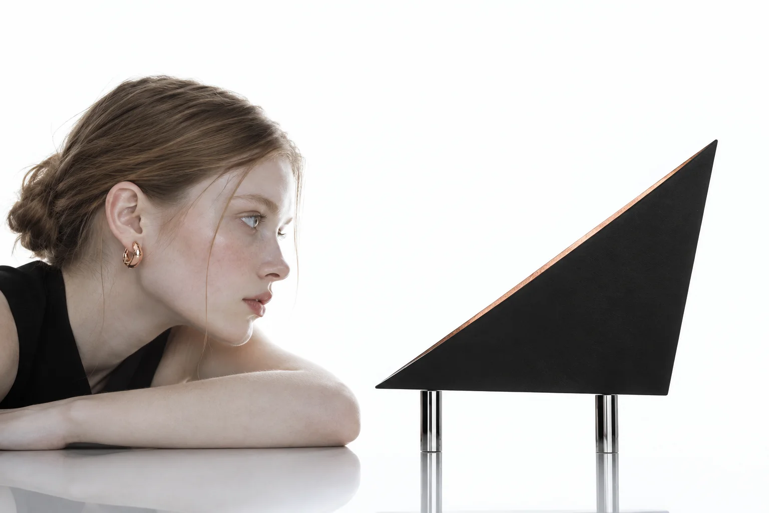 A woman in profile leans on a reflective white surface, looking intently at a minimalist black geometric sculpture in clean studio light.