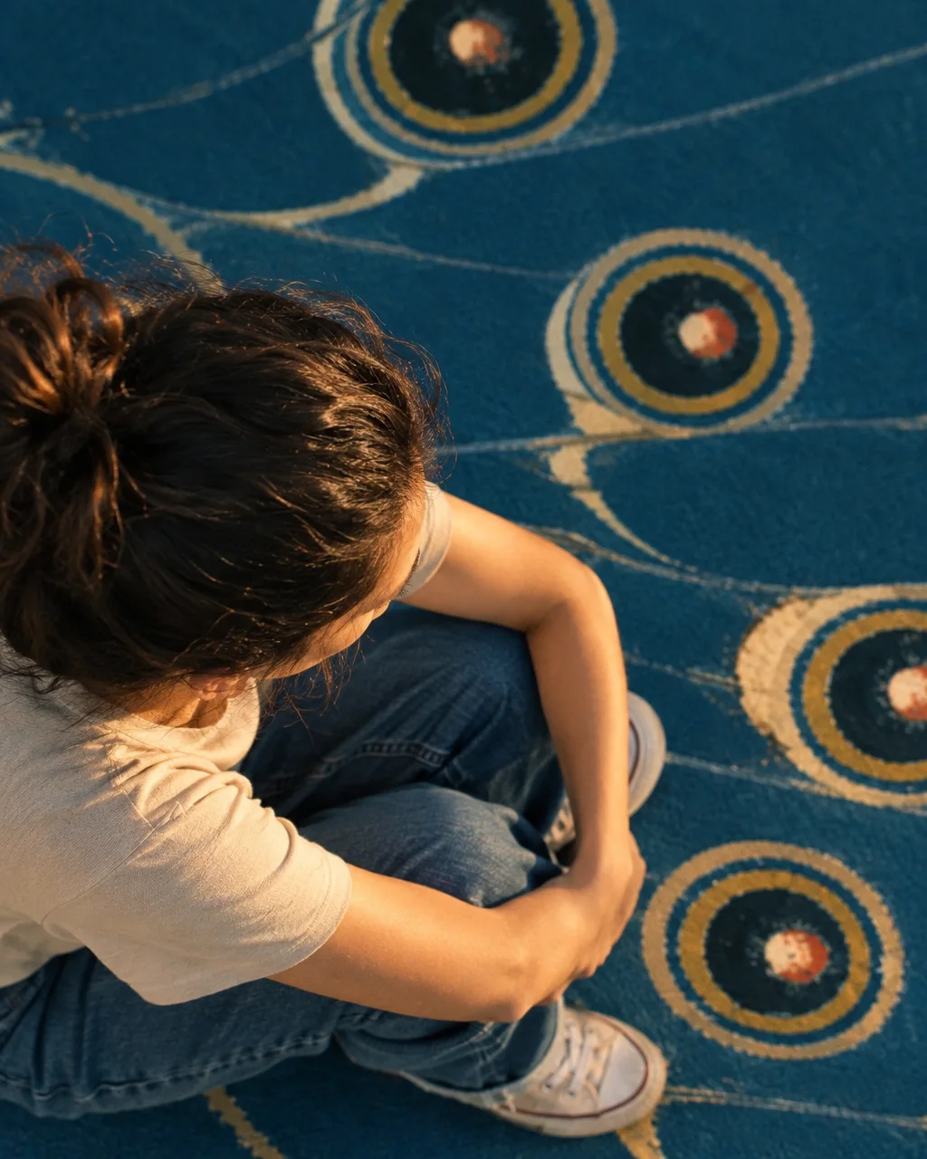A high-angle view of a woman hugging her knees while sitting on a patterned blue carpet in warm sunlight.