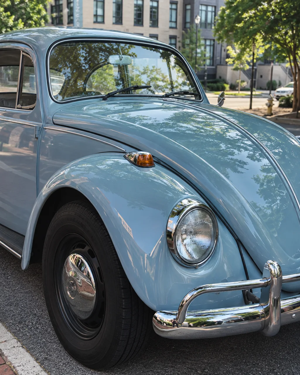 The front fender, hood, and chrome headlight of a classic light blue compact car parked along a sunlit city street.