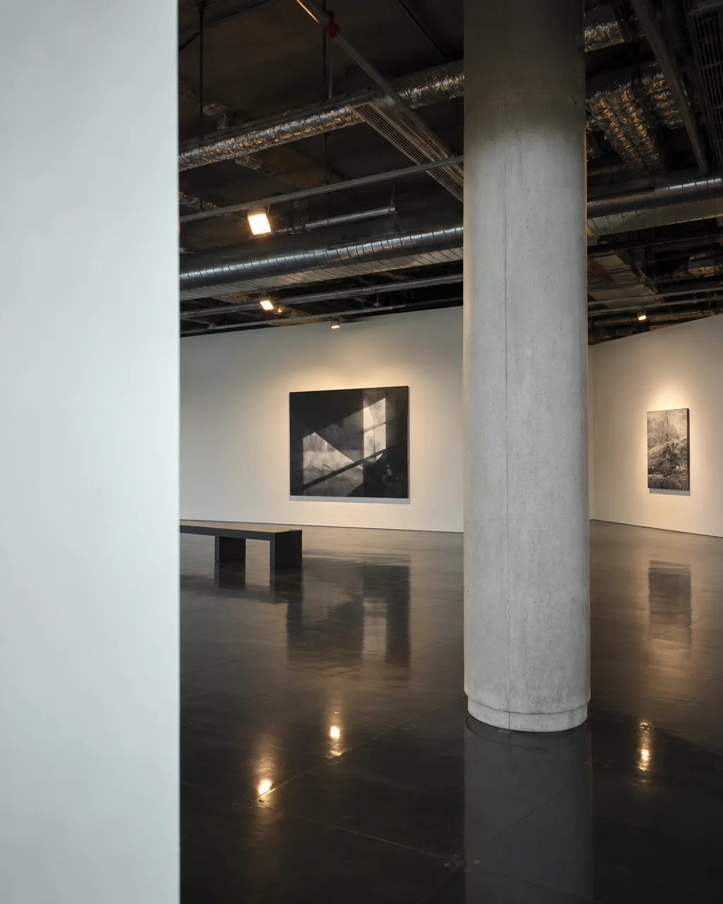 A modern gallery space with exposed industrial ceilings, polished reflective floors, and framed artwork hanging on white partition walls.