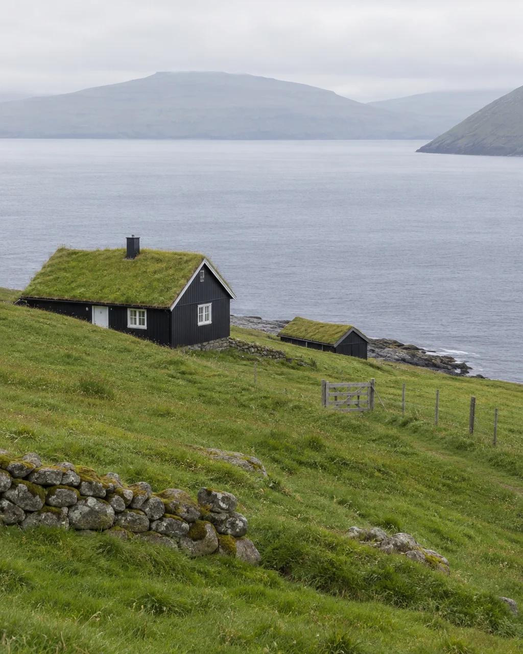 A black wooden cottage with a turf roof sits on a green coastal hillside overlooking a calm, misty sea and distant mountains.