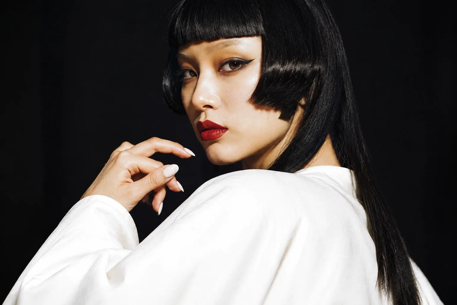 A woman with a sharp black hairstyle and bold red lipstick glances over her shoulder against a dark studio backdrop.