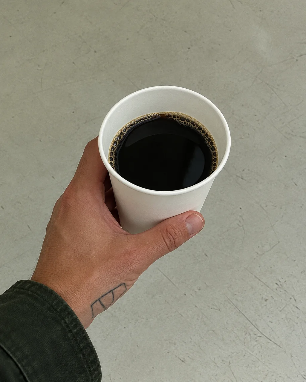 A hand holding a white paper cup filled with black coffee against a smooth, pale gray floor.