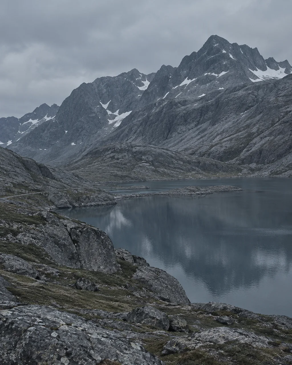 A calm mountain lake sits quietly beneath rugged, snow-dusted peaks under a heavy overcast sky.