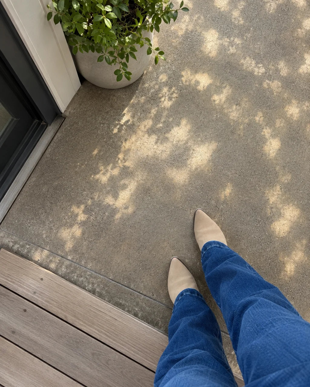 A top-down view of someone wearing beige pointed boots and blue jeans standing on a concrete patio in dappled sunlight.