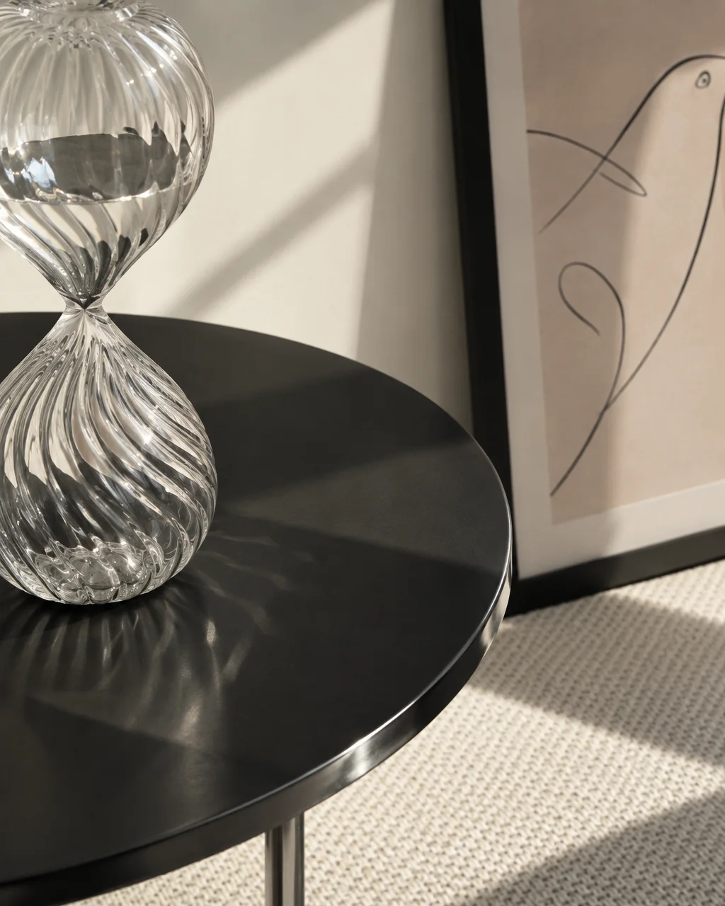 A ribbed glass hourglass rests on a sleek black table, casting refracted sunlight patterns beside framed artwork in a minimalist room.