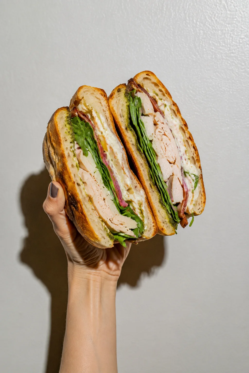 A hand holds up two halves of an artisan sandwich layered with sliced turkey, pesto, and greens against a pale wall.