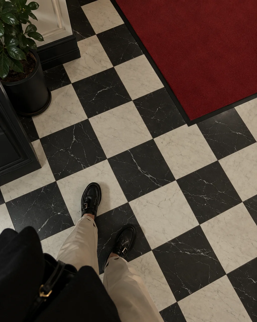 A downward point of view of black leather shoes on a classic black and white checkered marble floor beside a red runner.