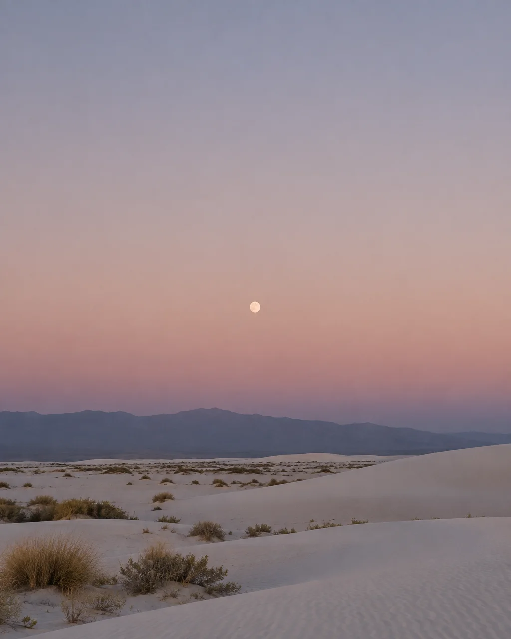 A full moon rises in a pastel gradient twilight sky above soft white sand dunes and distant mountain ranges.