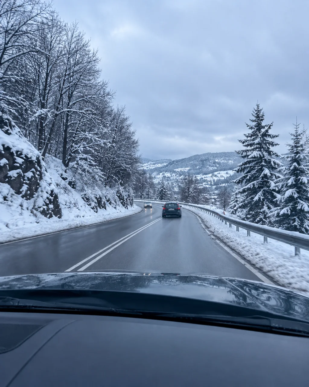 A wet asphalt road winds through snow-covered forest hills under an overcast sky, captured from behind a car windshield.