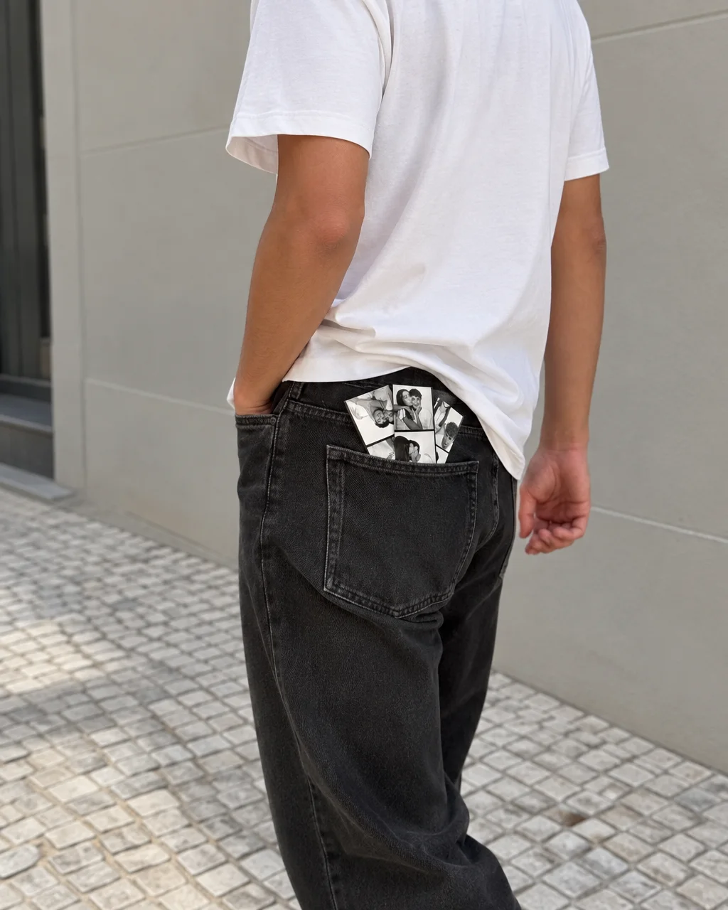 A person in a white t-shirt and dark jeans carries a black-and-white photobooth strip tucked into their back pocket on a paved street.